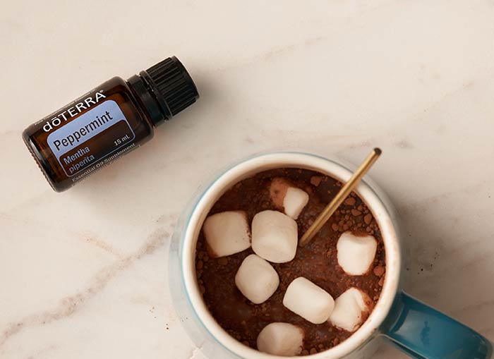 doTERRA Peppermint essential oil lying flat on a countertop. A mug, filled with hot chocolate, marshmallows, and a spoon, sit beside it. 