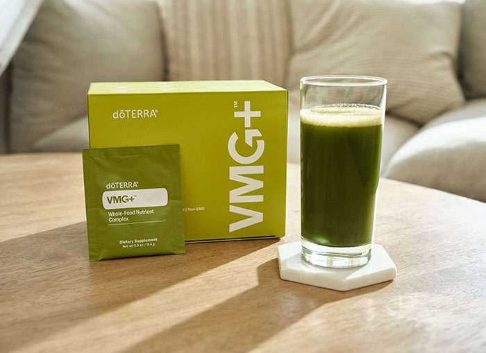 VMG+ box with a VMG+ sachet leaning against it sitting on a soft, wooden coffee table. A clear drinking glass on a white coaster, filled with VMG+, mixed with water, sits beside the box. A couch in the background with two pillows. 