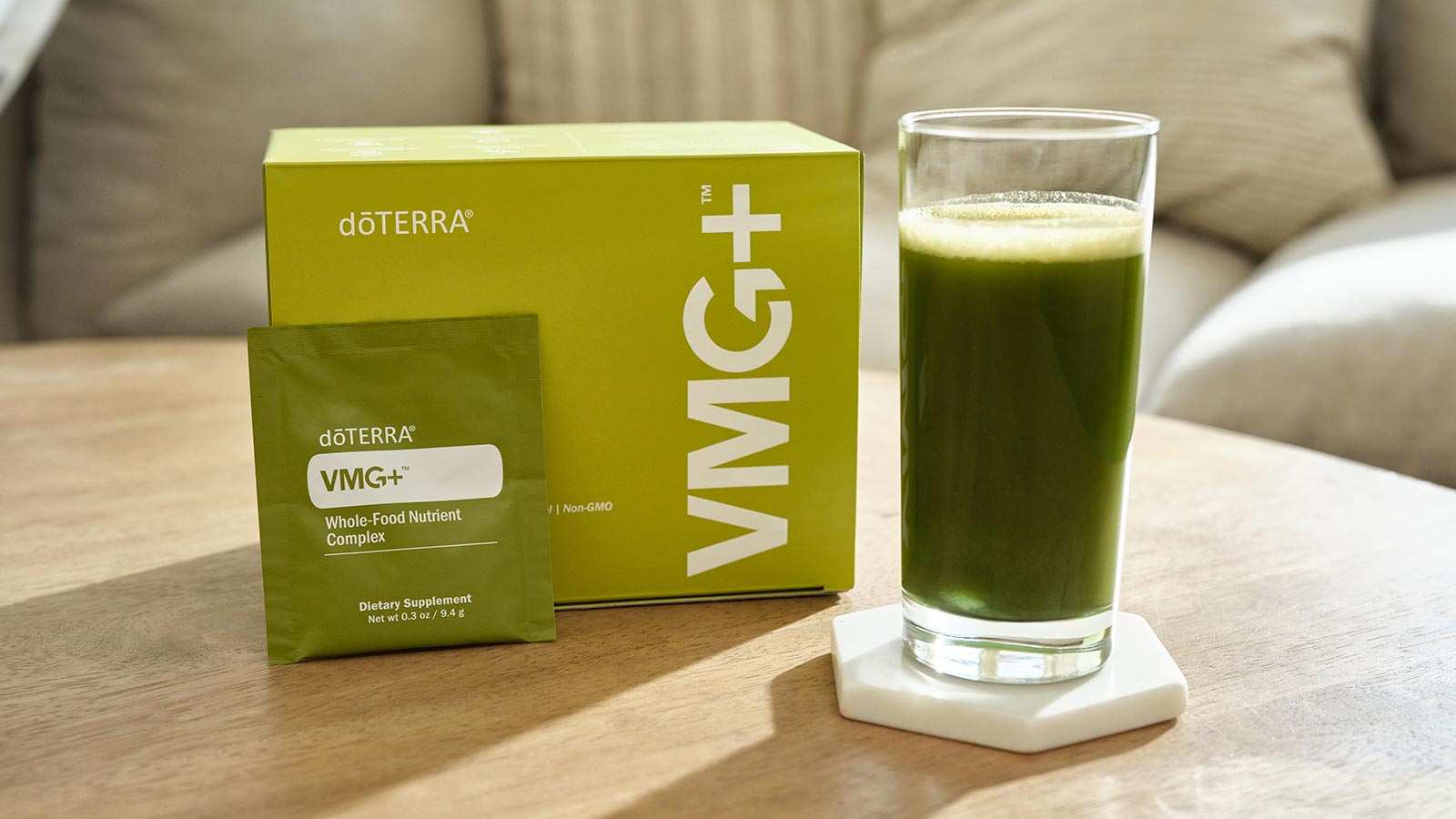 VMG+ box with a VMG+ sachet leaning against it sitting on a soft, wooden coffee table. A clear drinking glass on a white coaster, filled with VMG+, mixed with water, sits beside the box. A couch in the background with two pillows. 