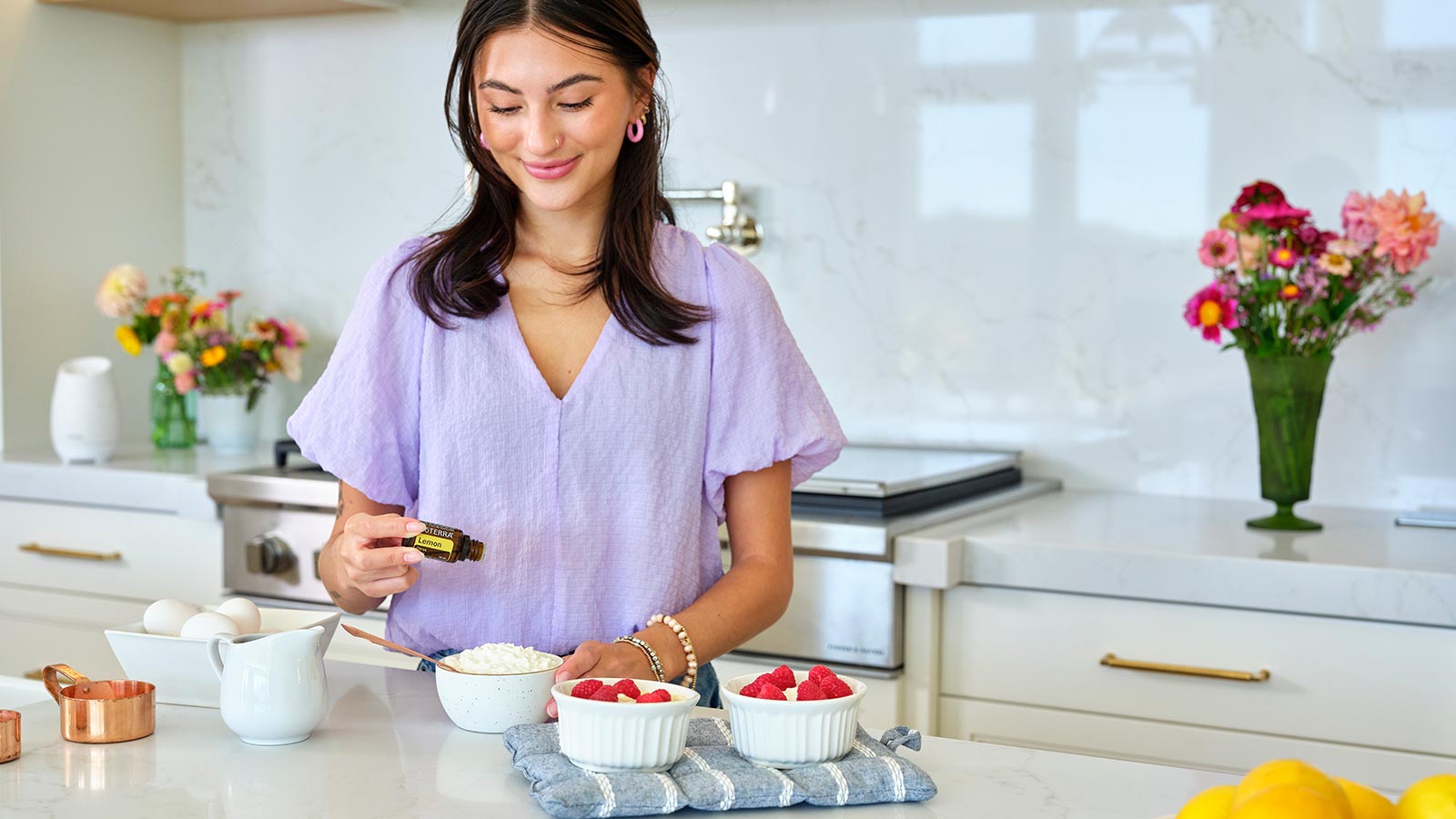 A woman standing in a primarily white kitchen making Lemon Protein Cheesecake. The ingredients and measuring tools surround her. Bouquets of flowers are behind her on the counter. 